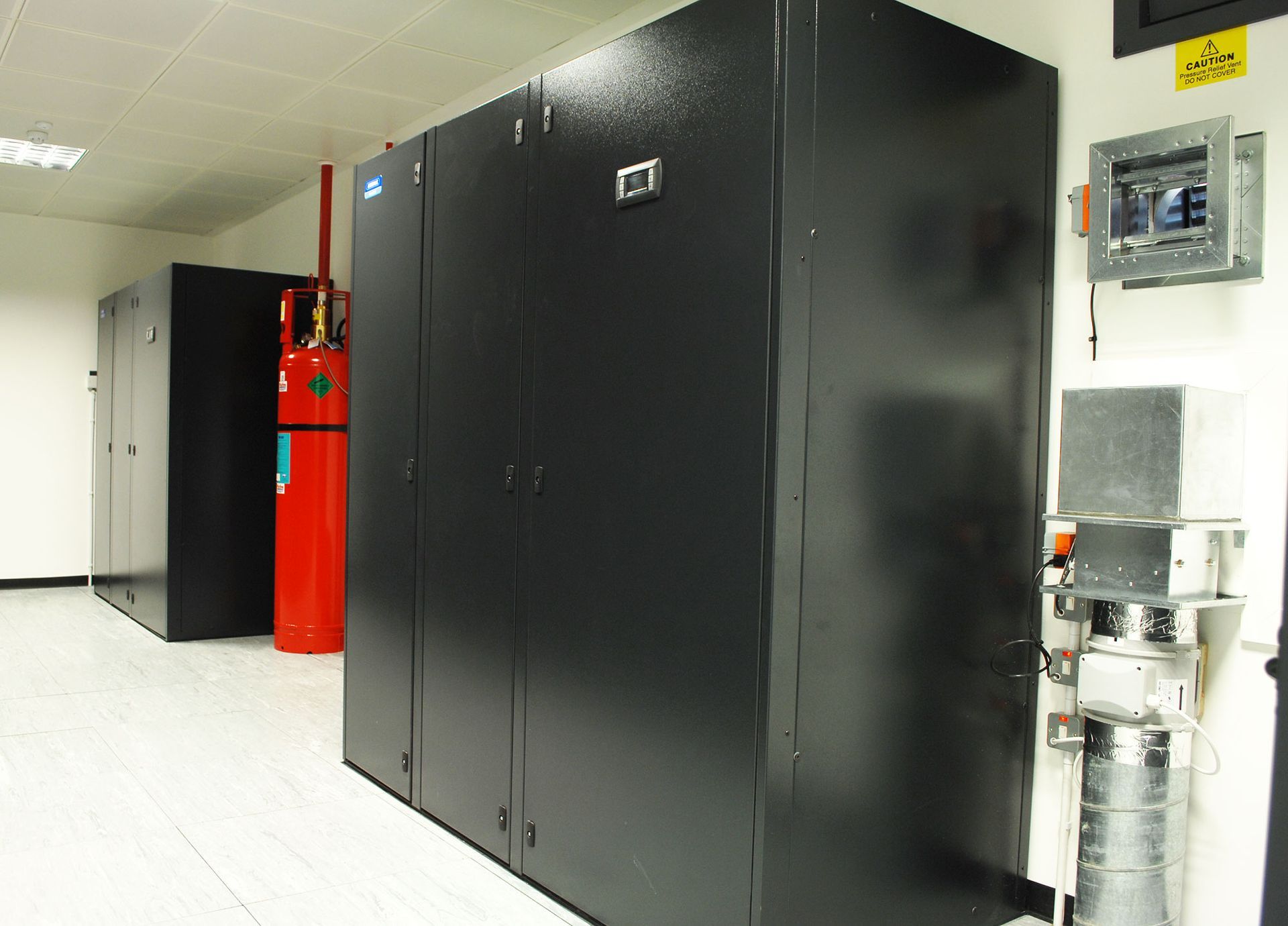 Row of large black server cabinets in a clean data center, with red equipment and wall-mounted controls on the right.
