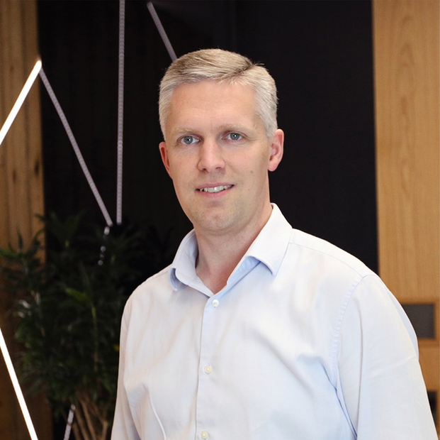 Portrait of a smiling man in a white shirt standing indoors against a modern office background