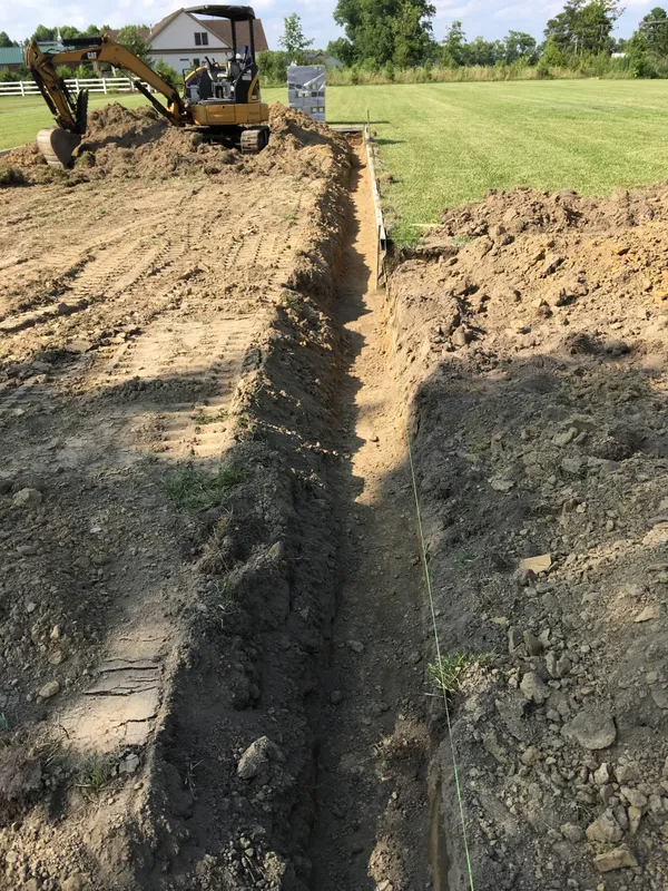 A backhoe digging a long trench in a dirt yard next to a grassy lawn.