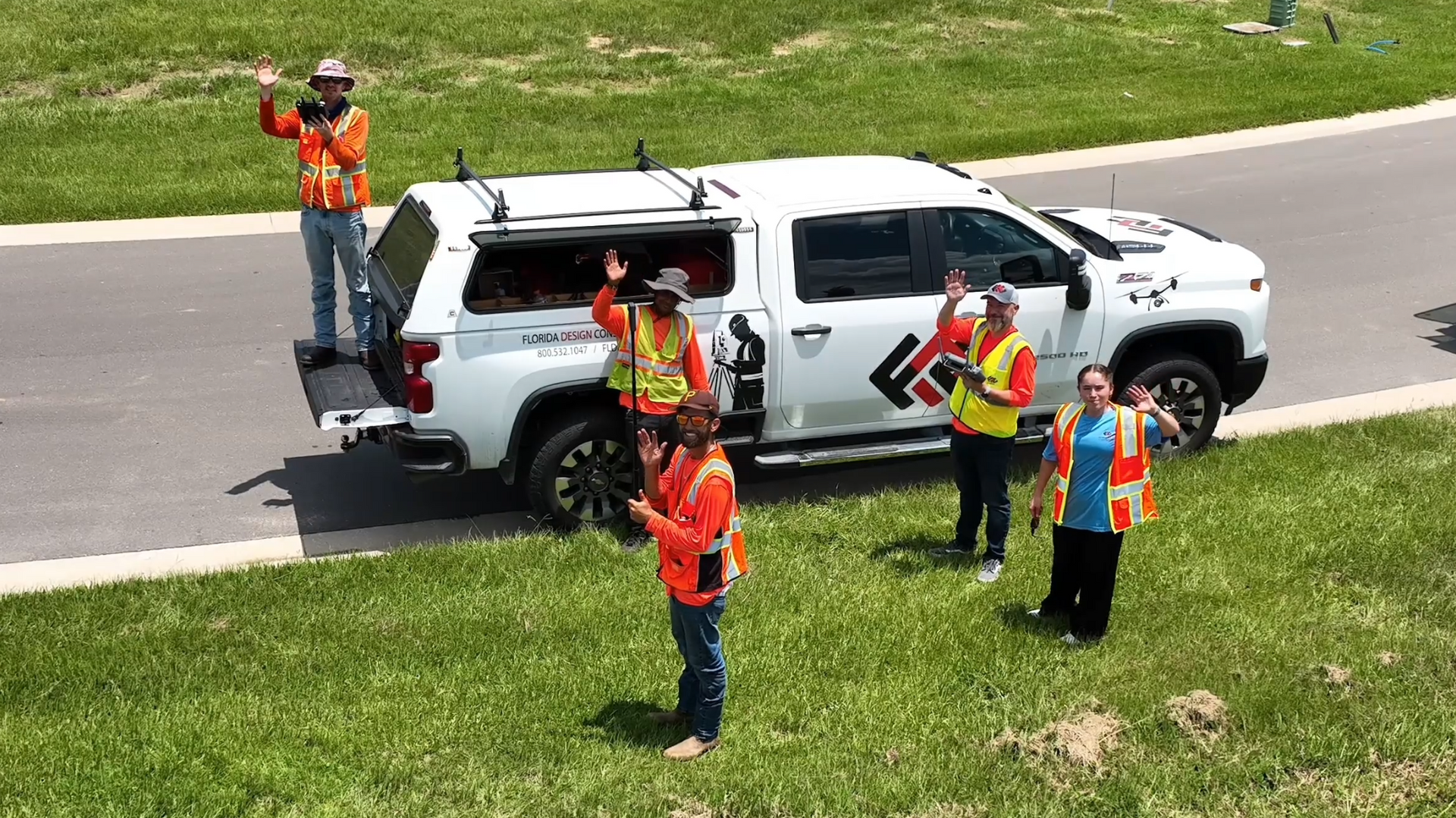 Construction workers by a white pickup truck on a grassy area. Several wear safety vests and wave.