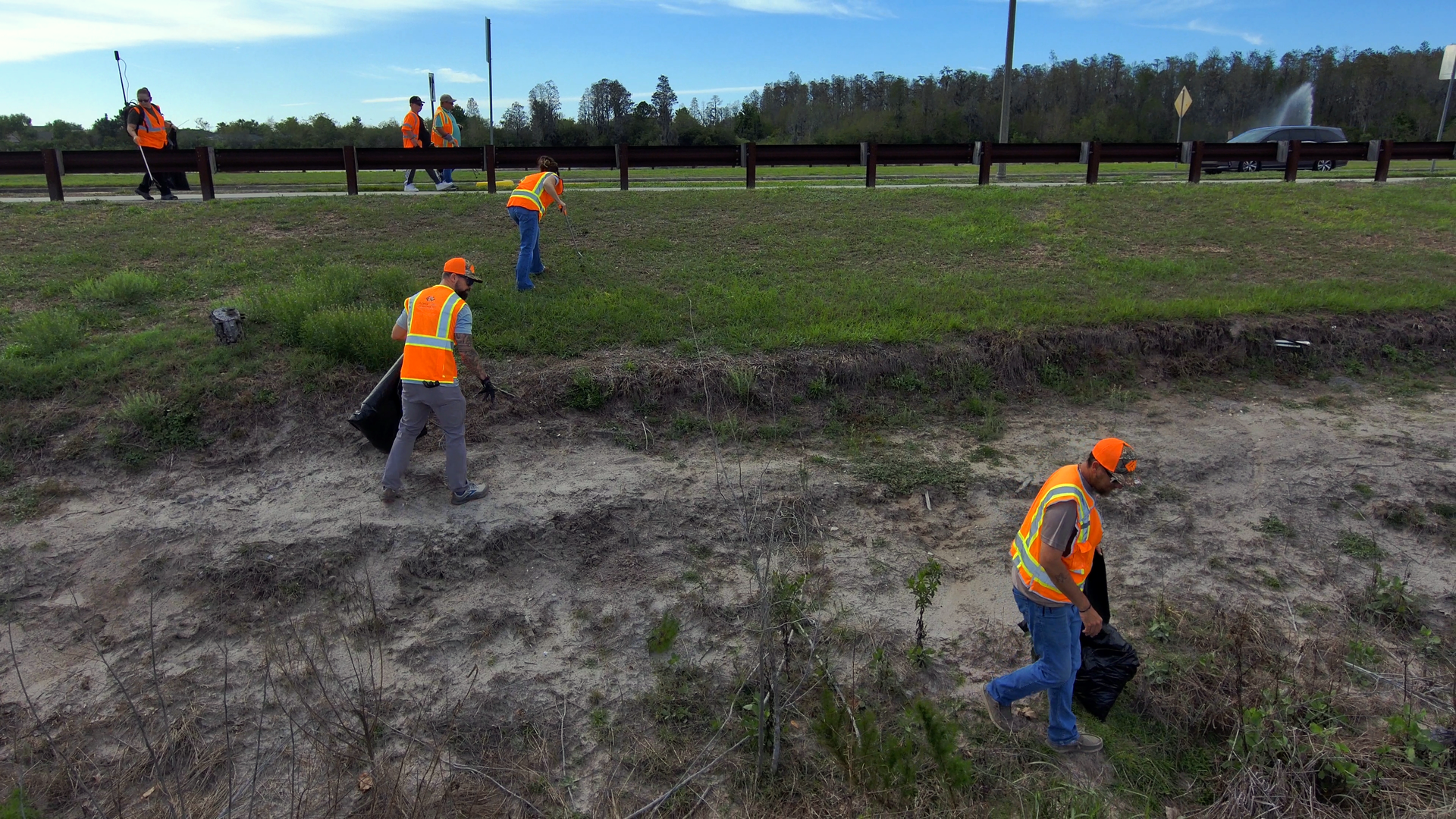 Workers in orange safety vests and hard hats clearing a muddy roadside ditch under an overcast sky