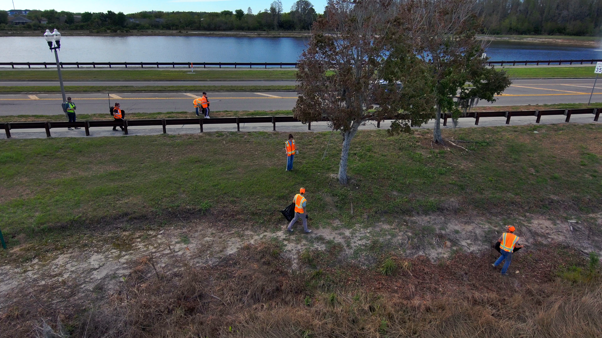 Workers in orange vests clearing brush along a roadside beside a river under trees