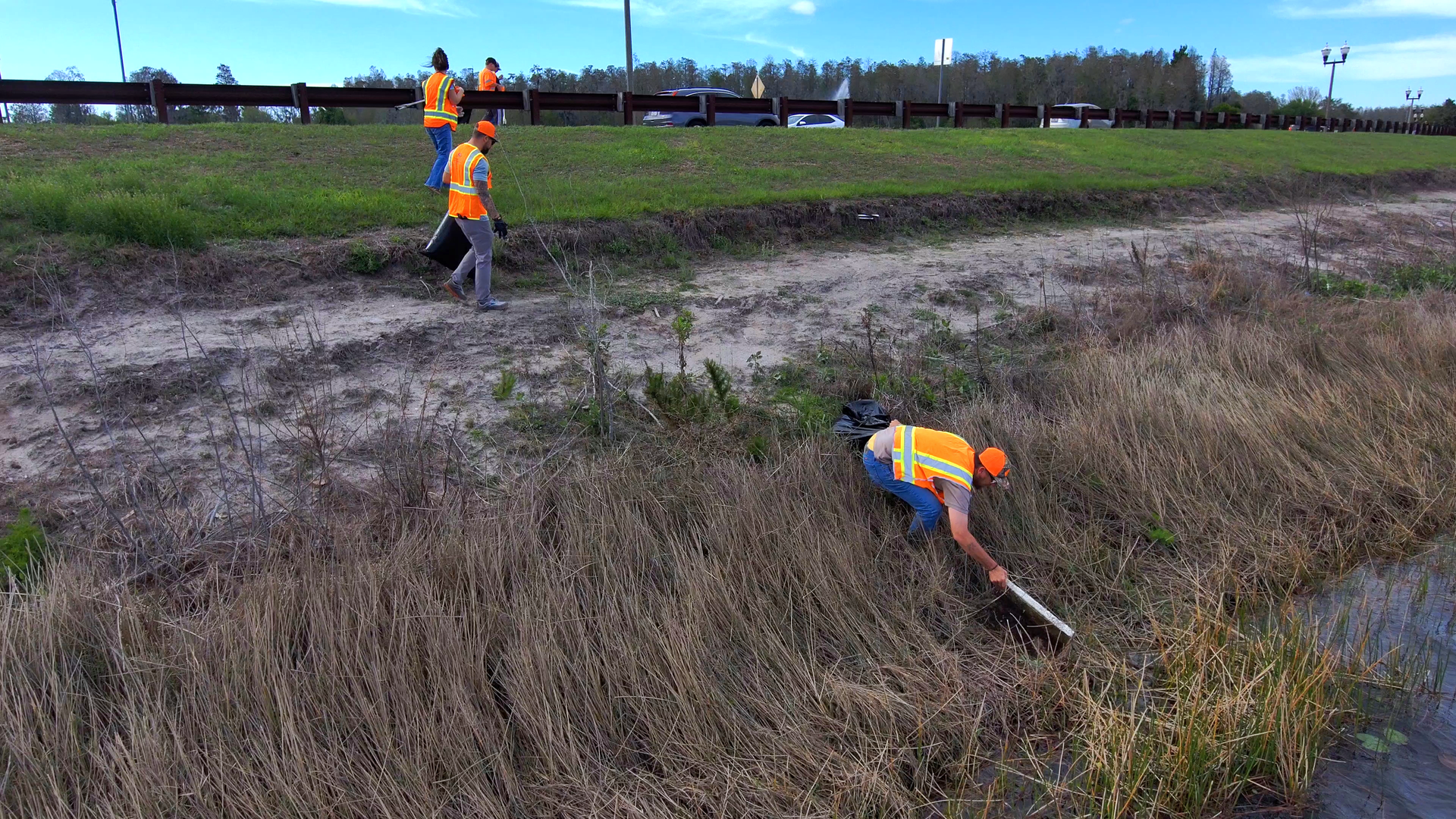 Workers in orange safety vests clearing brush along a grassy roadside ditch