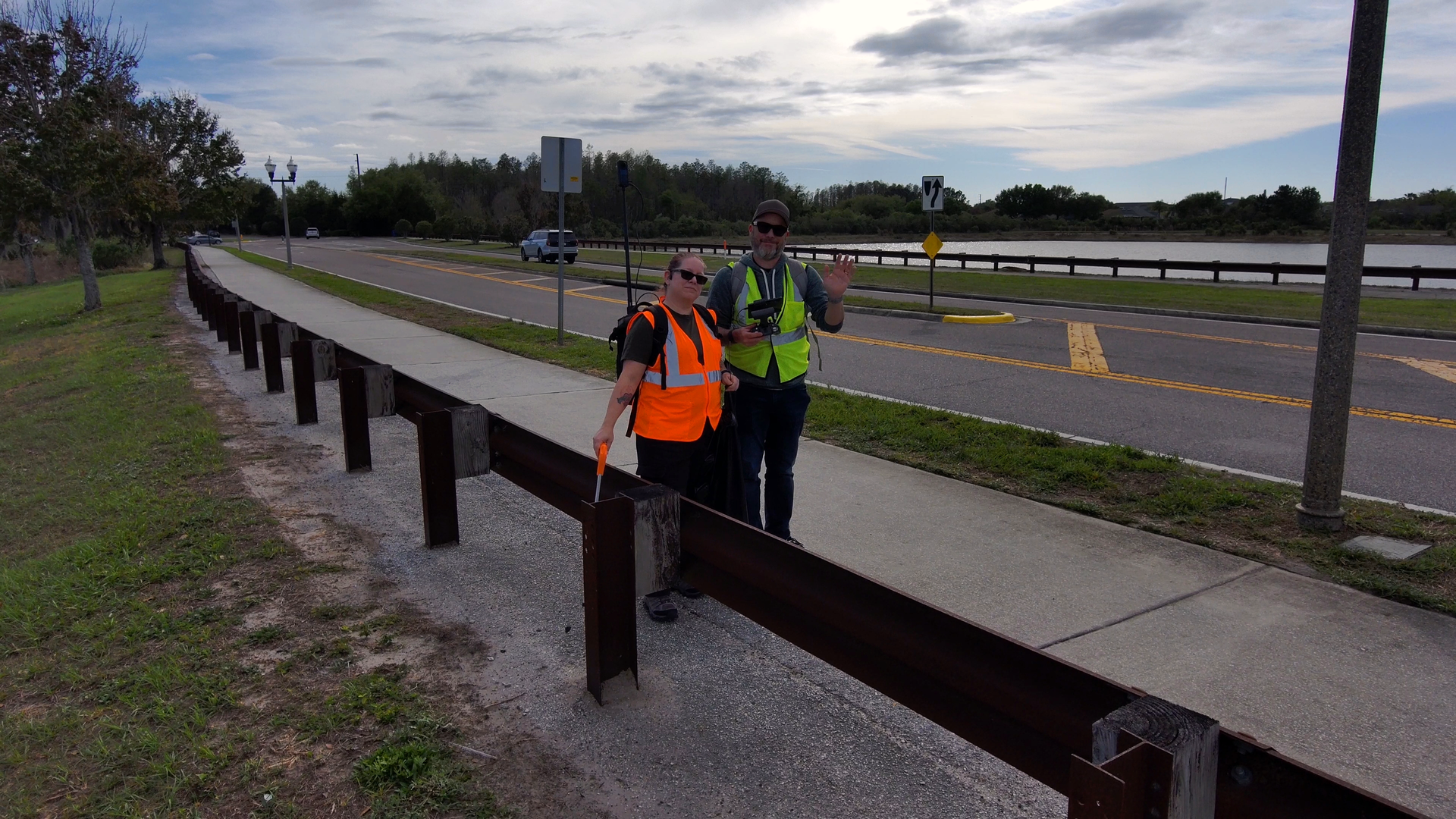 Two workers in safety vests stand by a roadside guardrail near a wet highway.