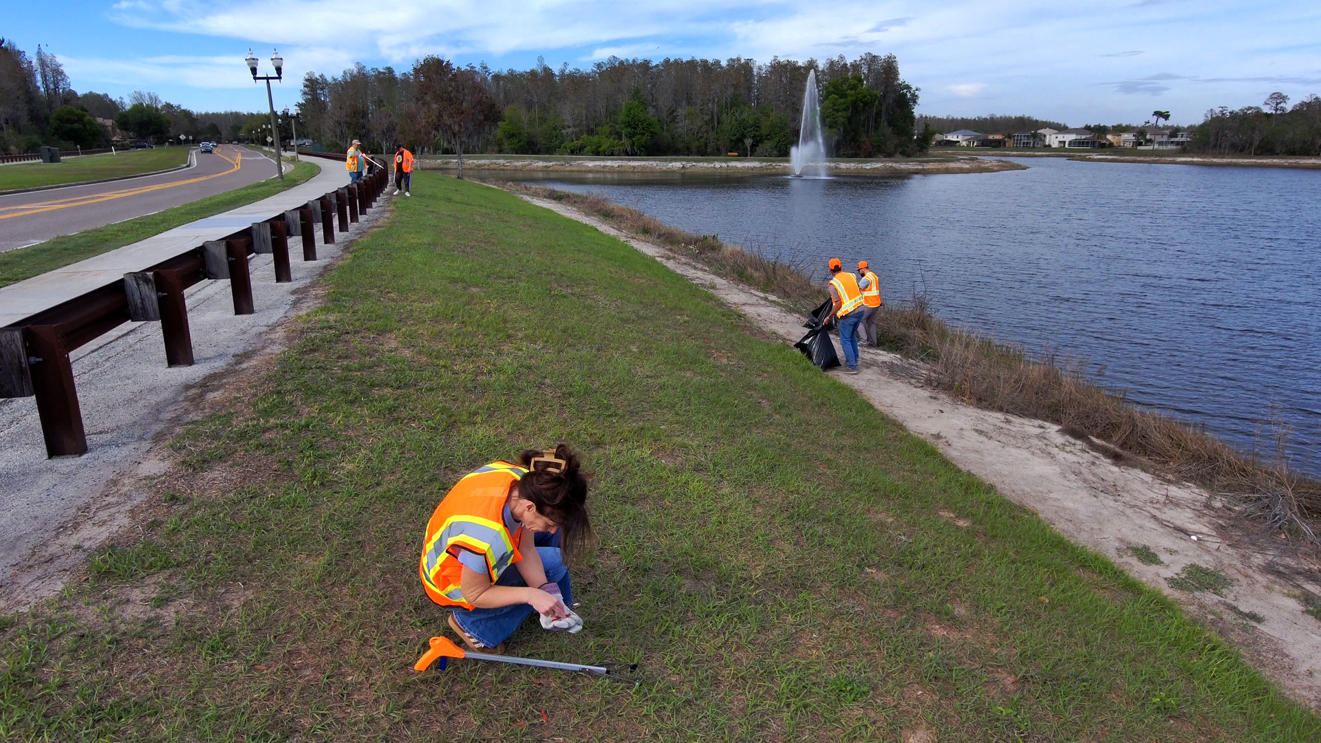 Children in orange safety vests planting along a grassy lakeside path near the water