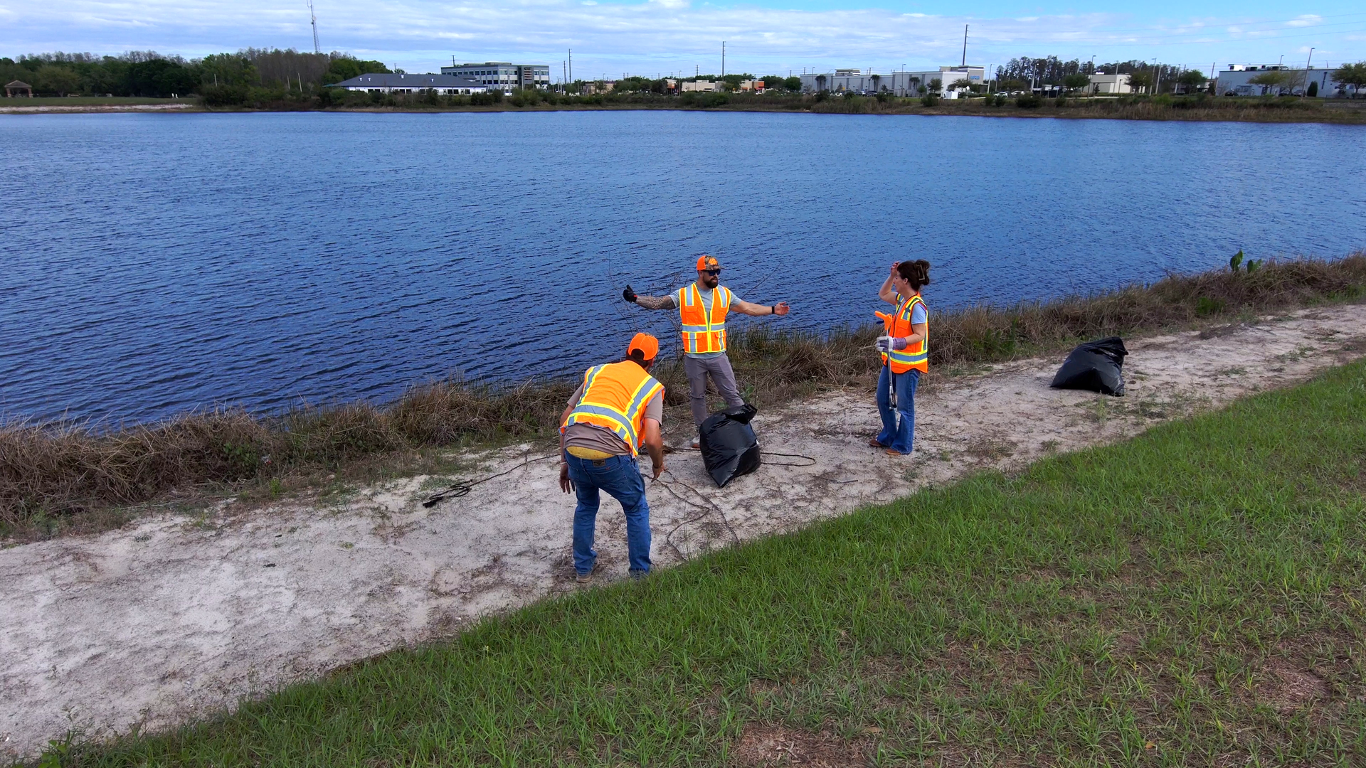 Workers in orange safety vests cleaning trash along a lakeshore beside a calm blue body of water
