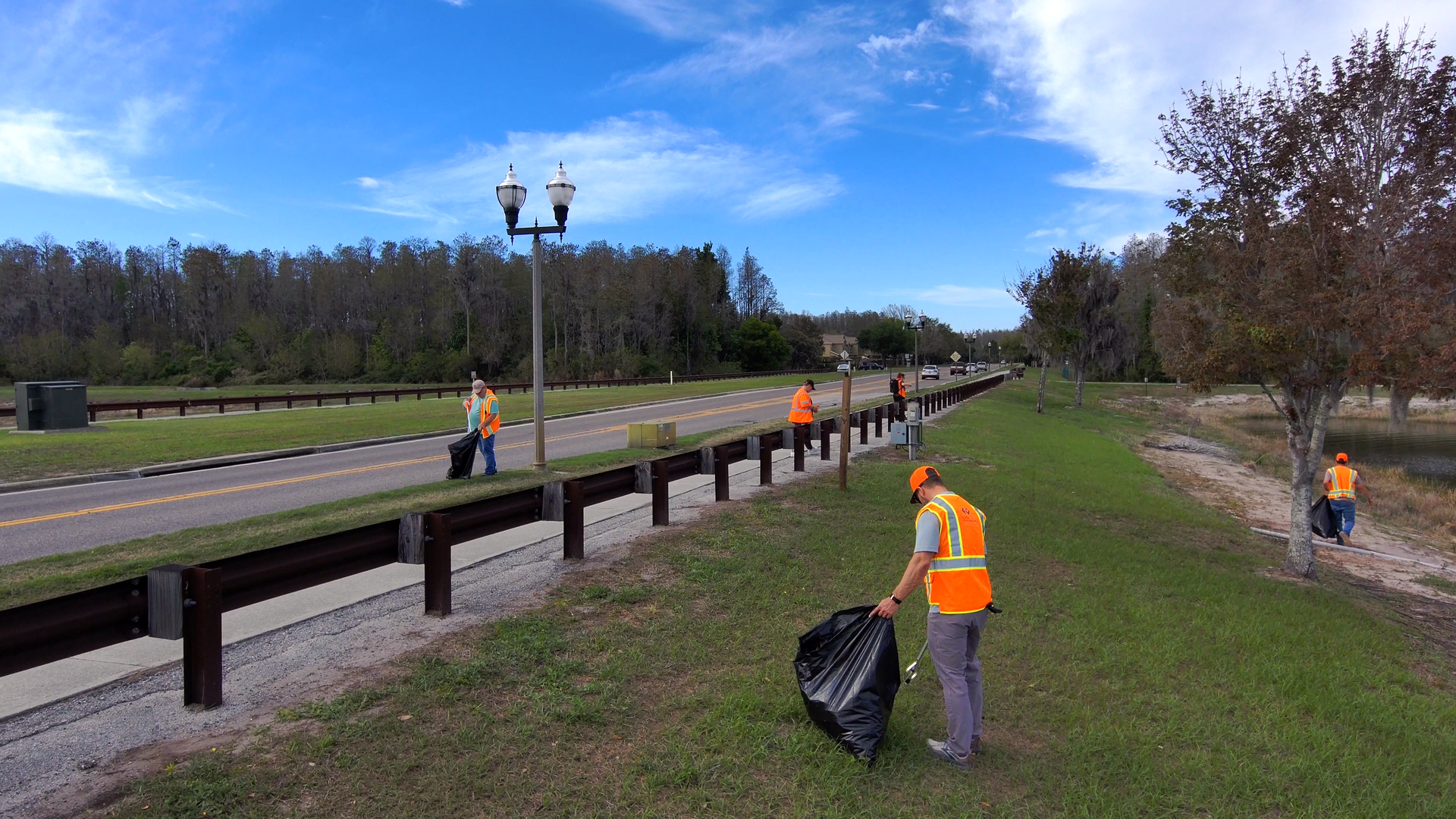 Workers in orange vests clean a roadside path beside a park and road under a blue sky.