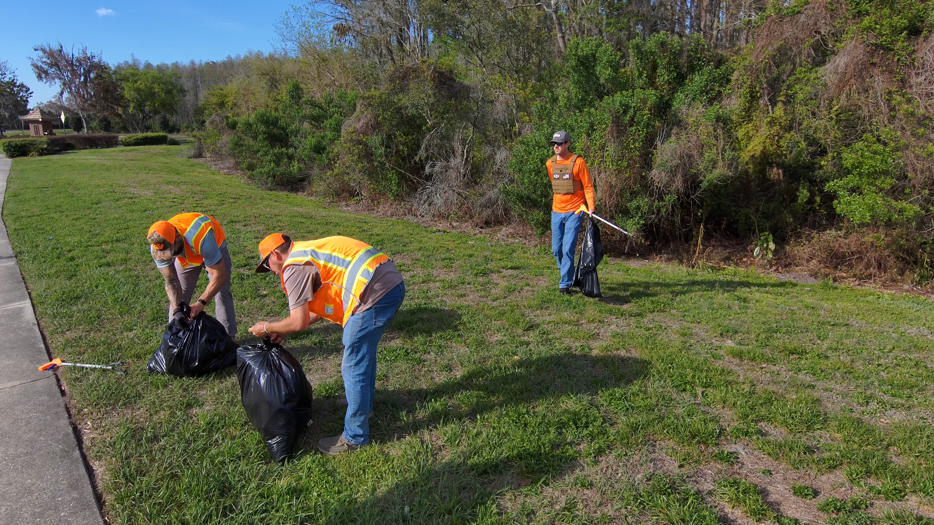 Workers in orange vests pick up trash along a grassy roadside path.