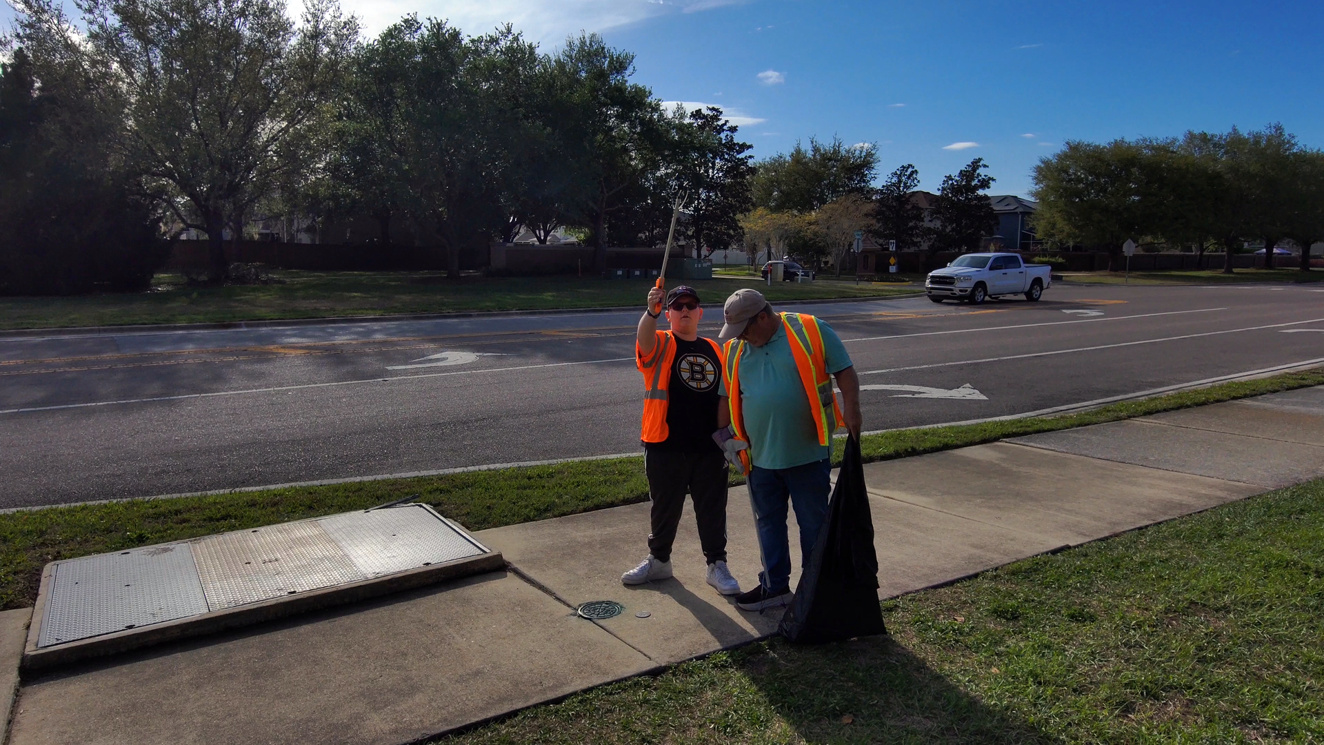 Two utility workers in orange safety vests inspect a roadside sidewalk near a street and parked car.