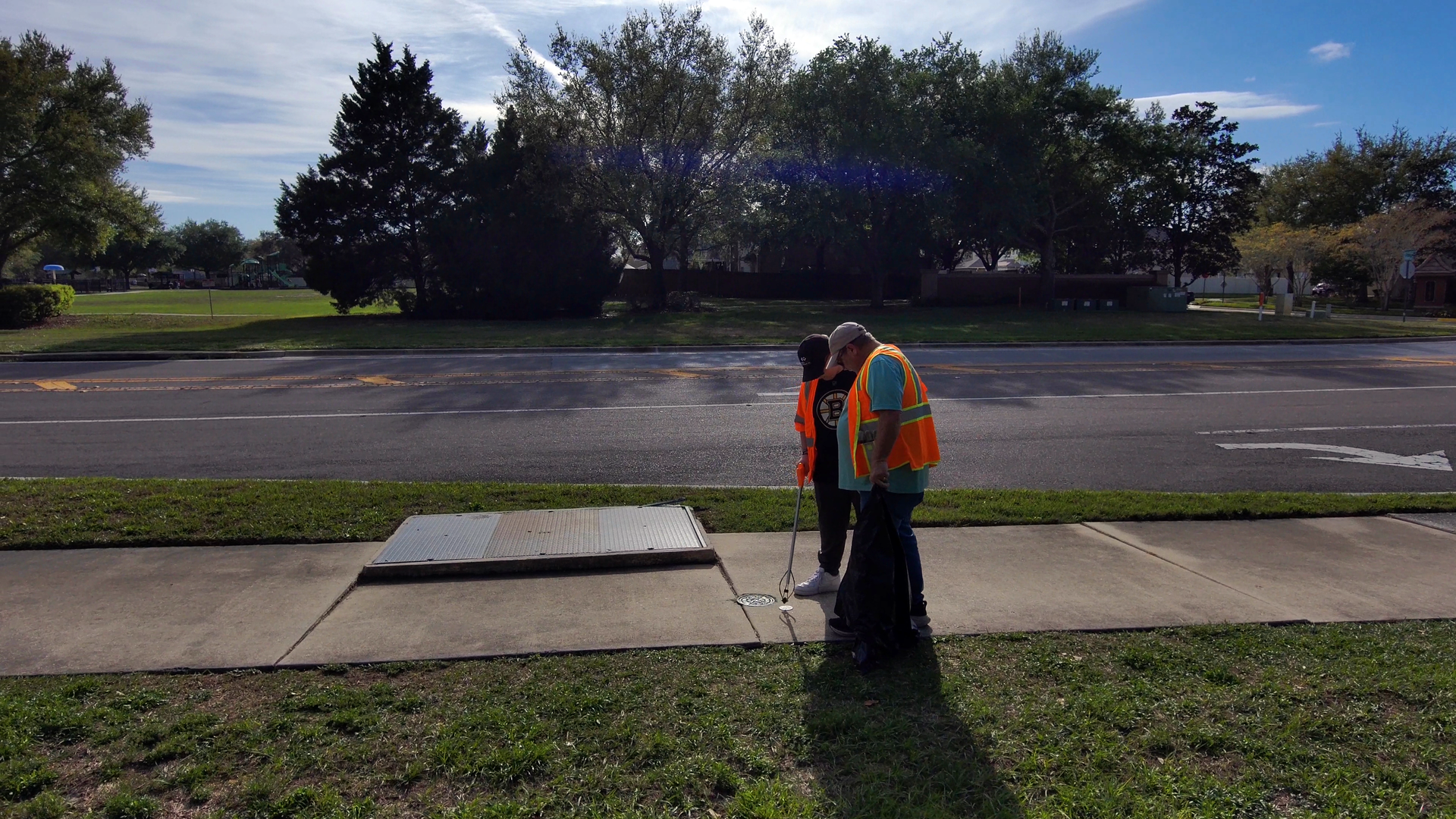 Two workers in orange safety vests inspect a sidewalk near a road on a sunny day.