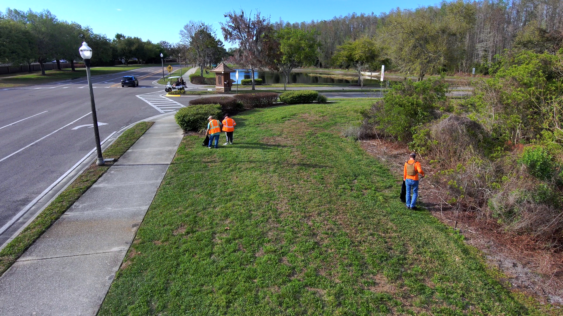 Workers in orange vests clearing brush along a grassy roadside beside a parking lot
