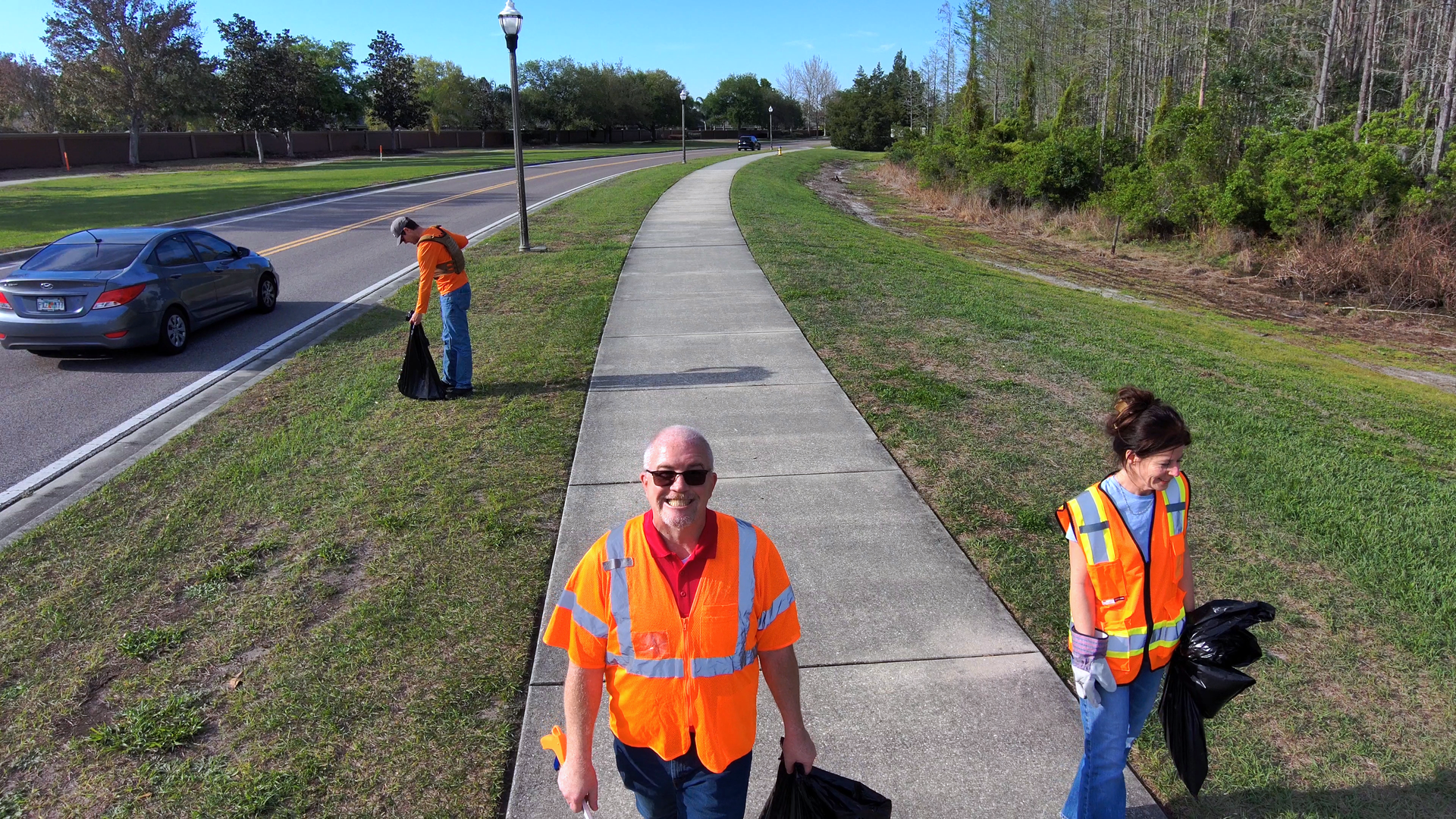 Three workers in orange safety vests on a roadside path, one carrying a tool bag.