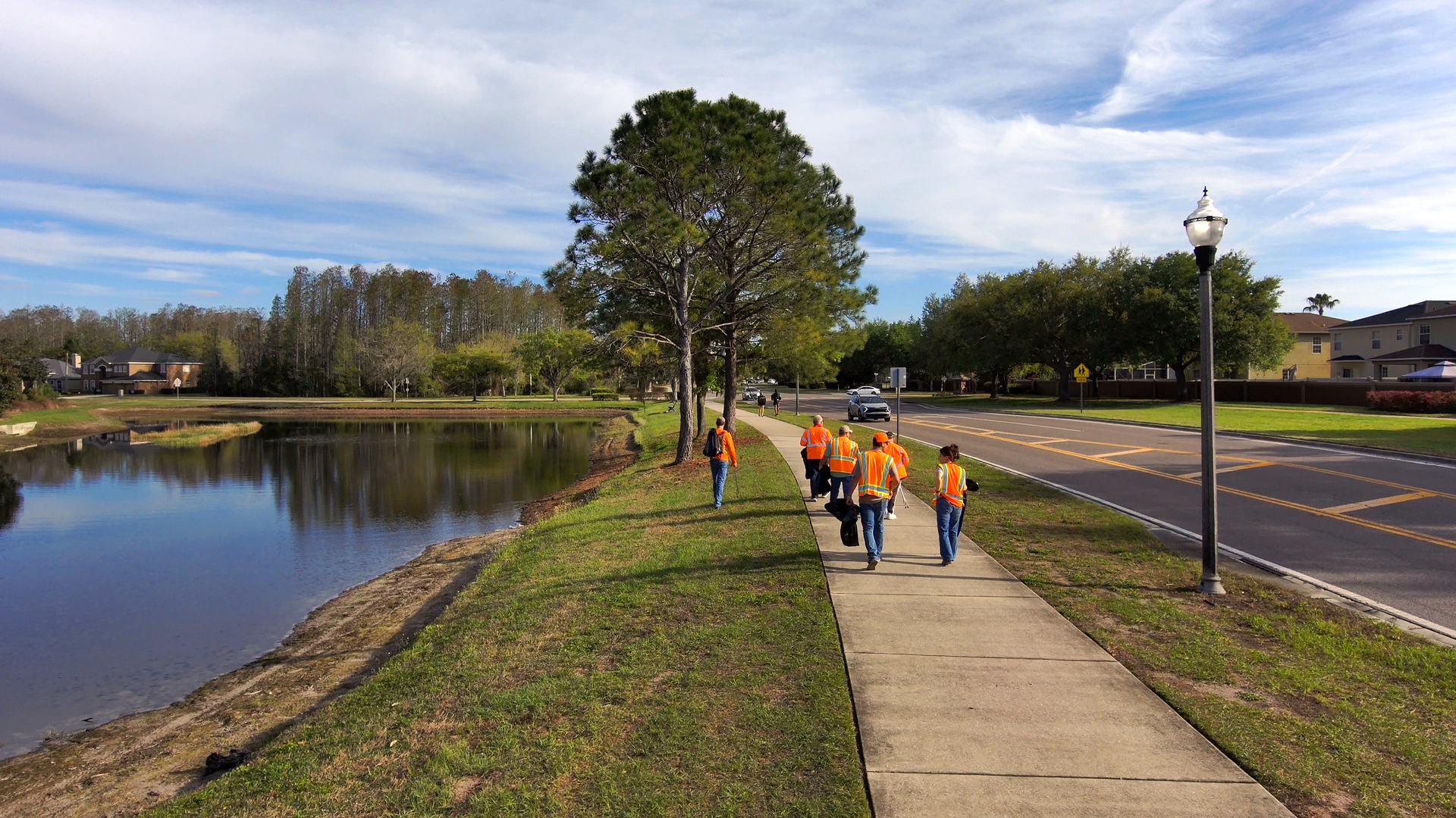 People in orange shirts walk along a path beside a lake and road under a partly cloudy sky