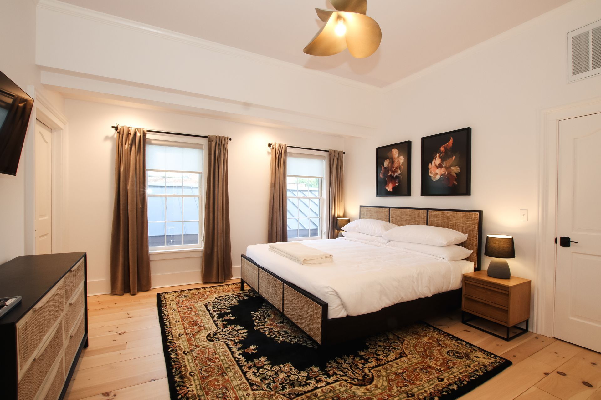 Picture of the room called Mason featuring a king bed with two windows flanked with long neutral colored drapes. Two flower themed pieces of art hang above the cane headboard and a tulip inspired light fixture is above the bed.