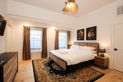 A large room with king size bed and two windows draped by neutral colored curtains. A tulip shaped pendant light hangs over the bed.
