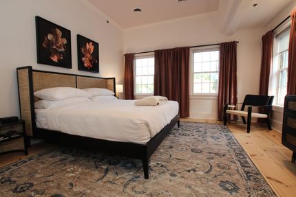 A large room with king size bed and four windows draped by pink curtains. A tulip shaped pendant light hangs over the bed.