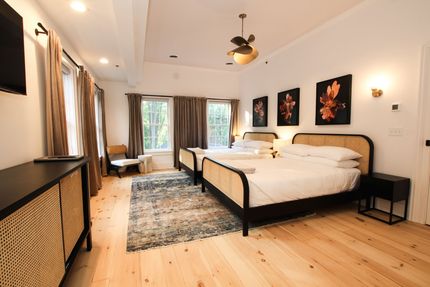 A large room with two queen size beds and four windows draped by neutral colored curtains. A tulip shaped pendant light hangs over the bed.