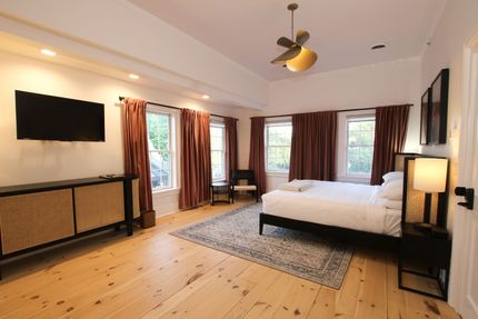 A large room with king size bed and four windows draped by pink curtains. A tulip shaped pendant light hangs over the bed.