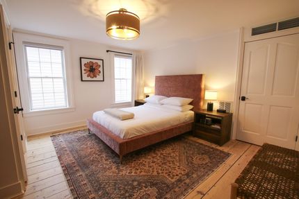 A medium size room with queen size bed and two windows. One window is draped by white velvet curtains. A pendant light hangs over the bed. A large pink headboard adorns the bed.