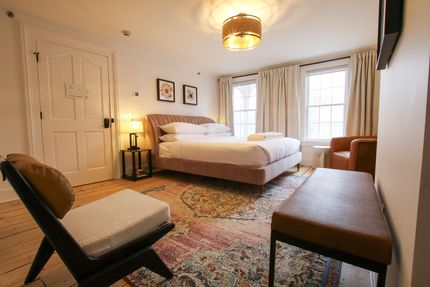 A large room with king size bed and two windows draped by white velvet curtains. Pink upholstery adorns the bed and carpet. A pendant light hangs over the bed.