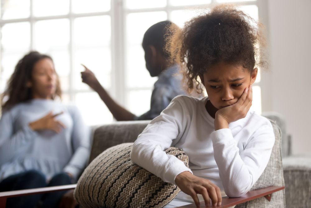 A little girl is sitting on a couch while her parents argue in the background.