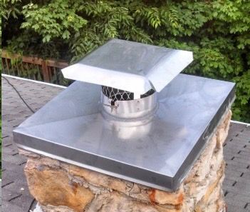 A metal chimney cap with a mesh screen sits atop a stone chimney on a roof with asphalt shingles.
