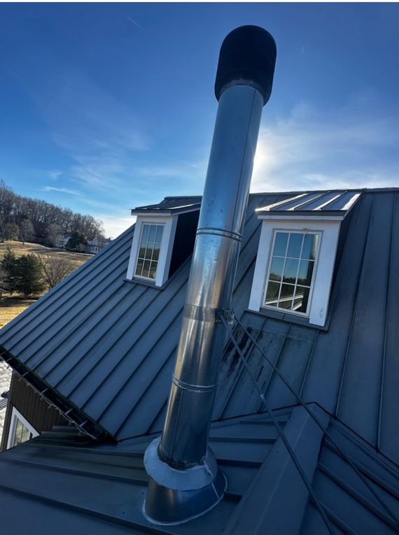 A tall, stainless steel chimney pipe extending from a dark metal roof, situated between two small dormer windows.