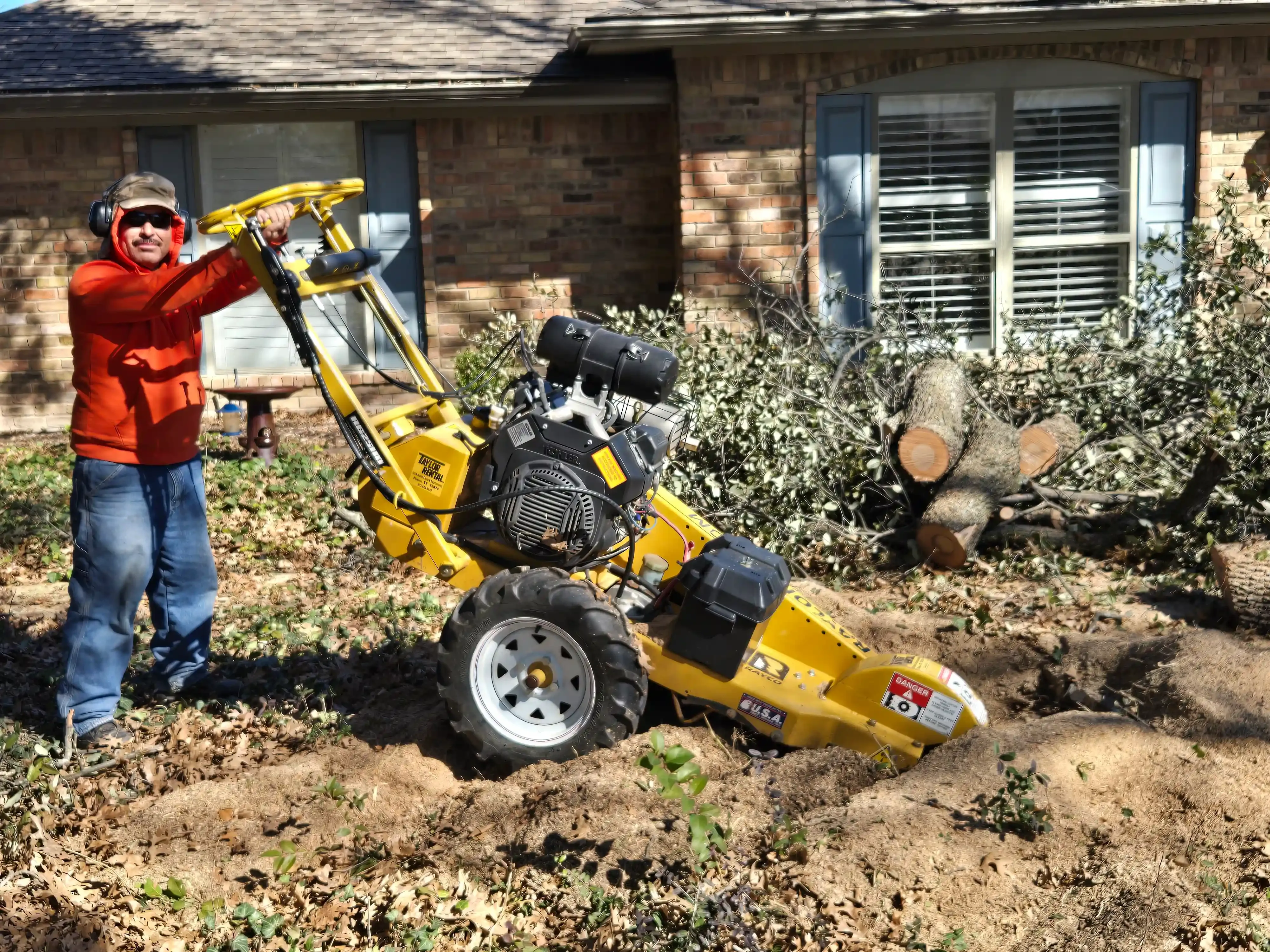 A man is standing next to a yellow stump grinder in front of a house.