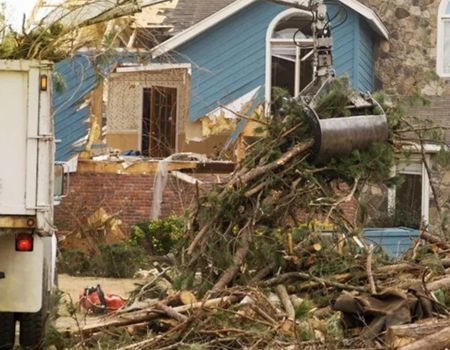 Storm-damaged blue house with fallen trees and debris blocking the yard