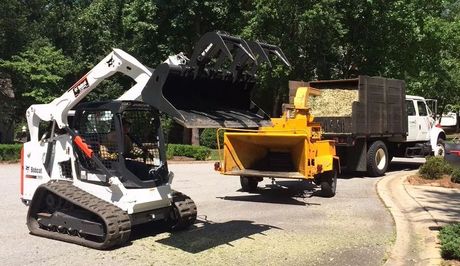White skid-steer loader pours black mulch into yellow wood chipper beside a dump truck on a tree-lined road