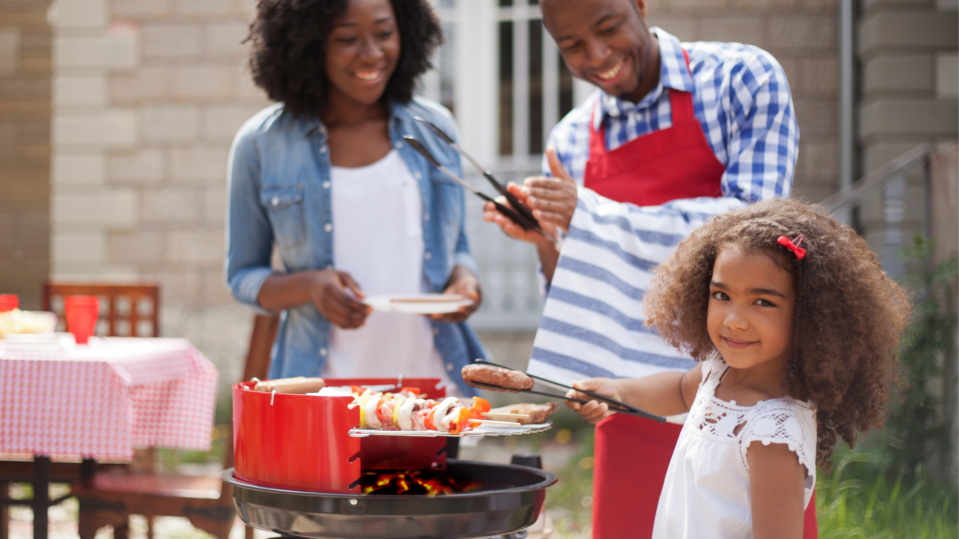 A family grilling outdoors on a sunny day, with a young child using tongs to flip skewers on the barbecue.