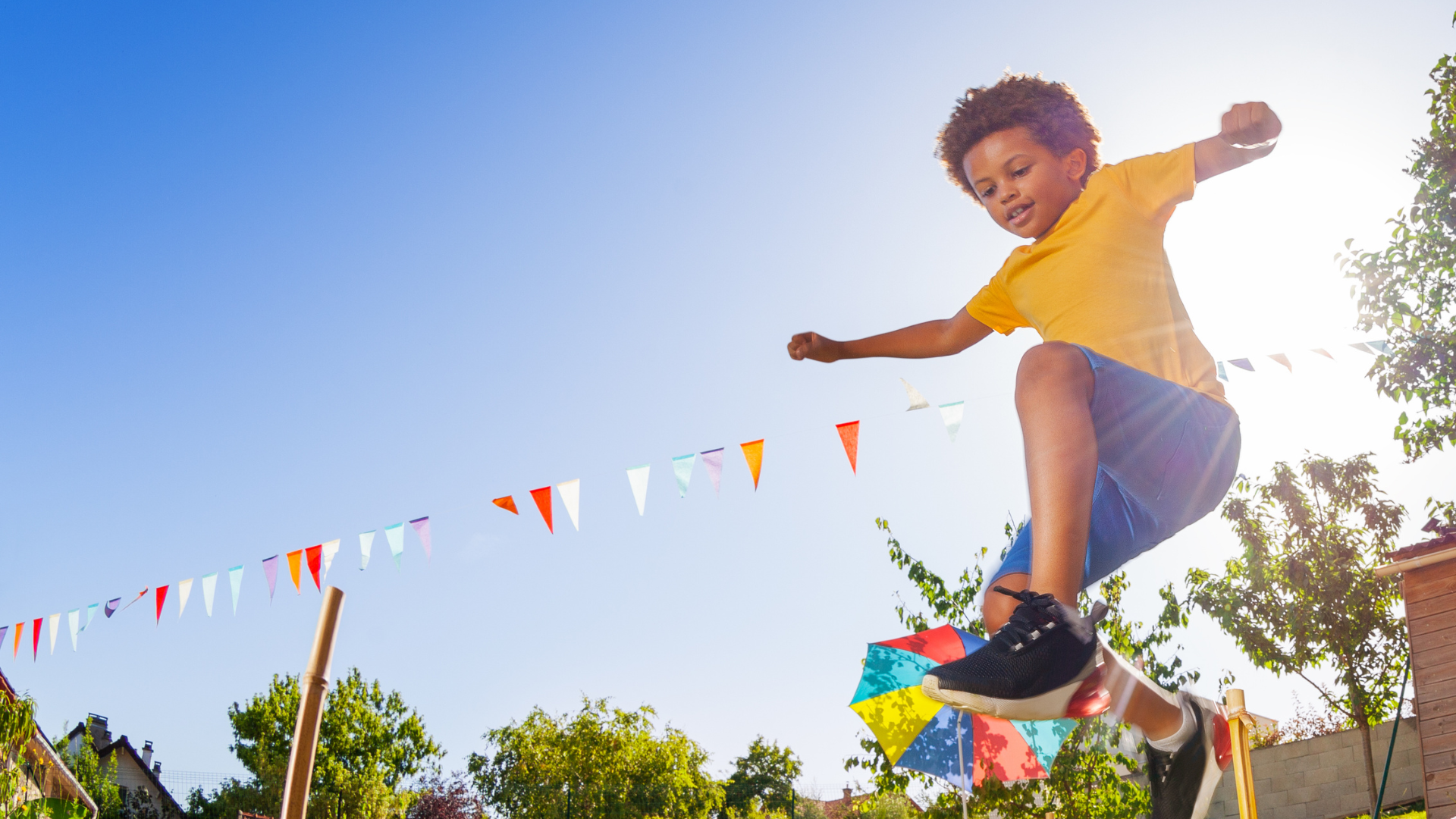 A person in a yellow shirt and blue shorts jumps outdoors against a bright blue sky with colorful bunting.