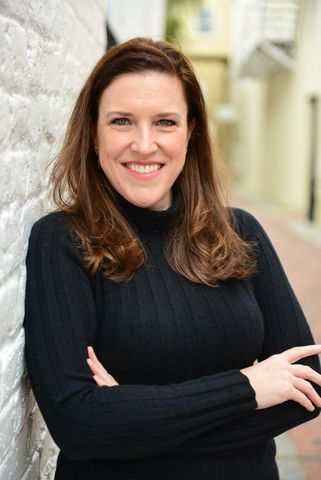 A woman in a black sweater is leaning against a white wall and smiling.