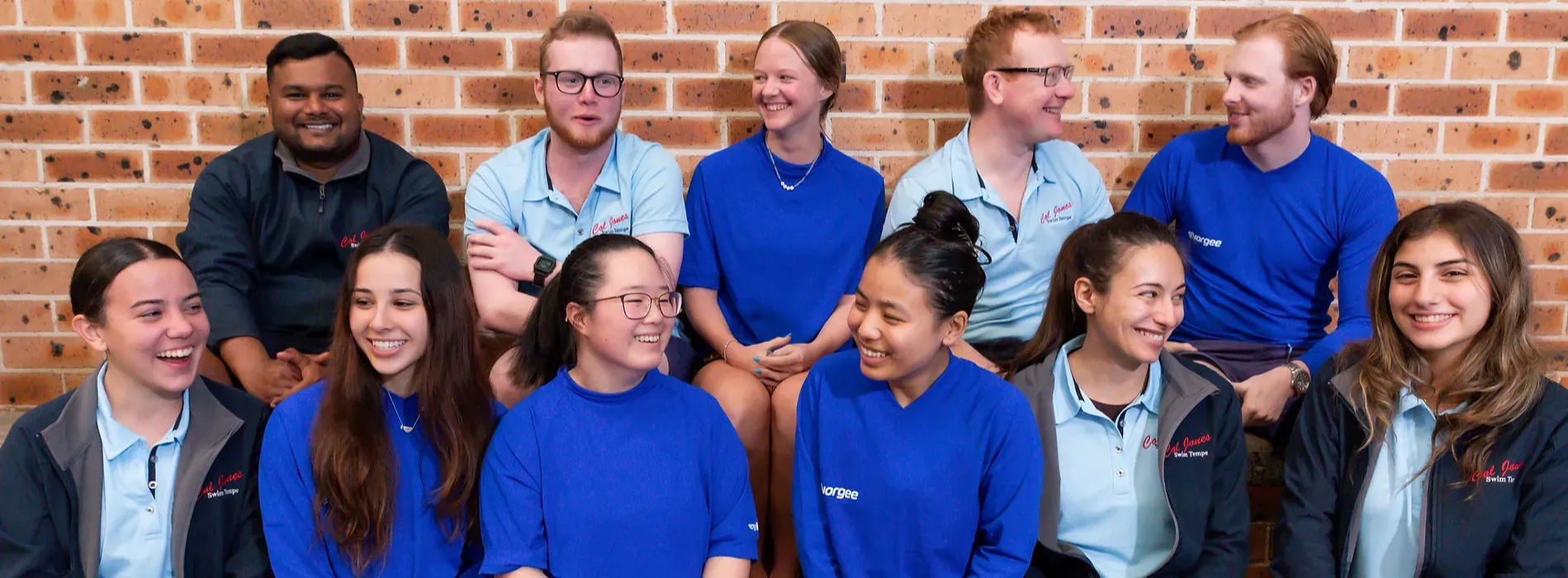 Group of smiling people in front of a brick wall, wearing blue shirts and jackets.