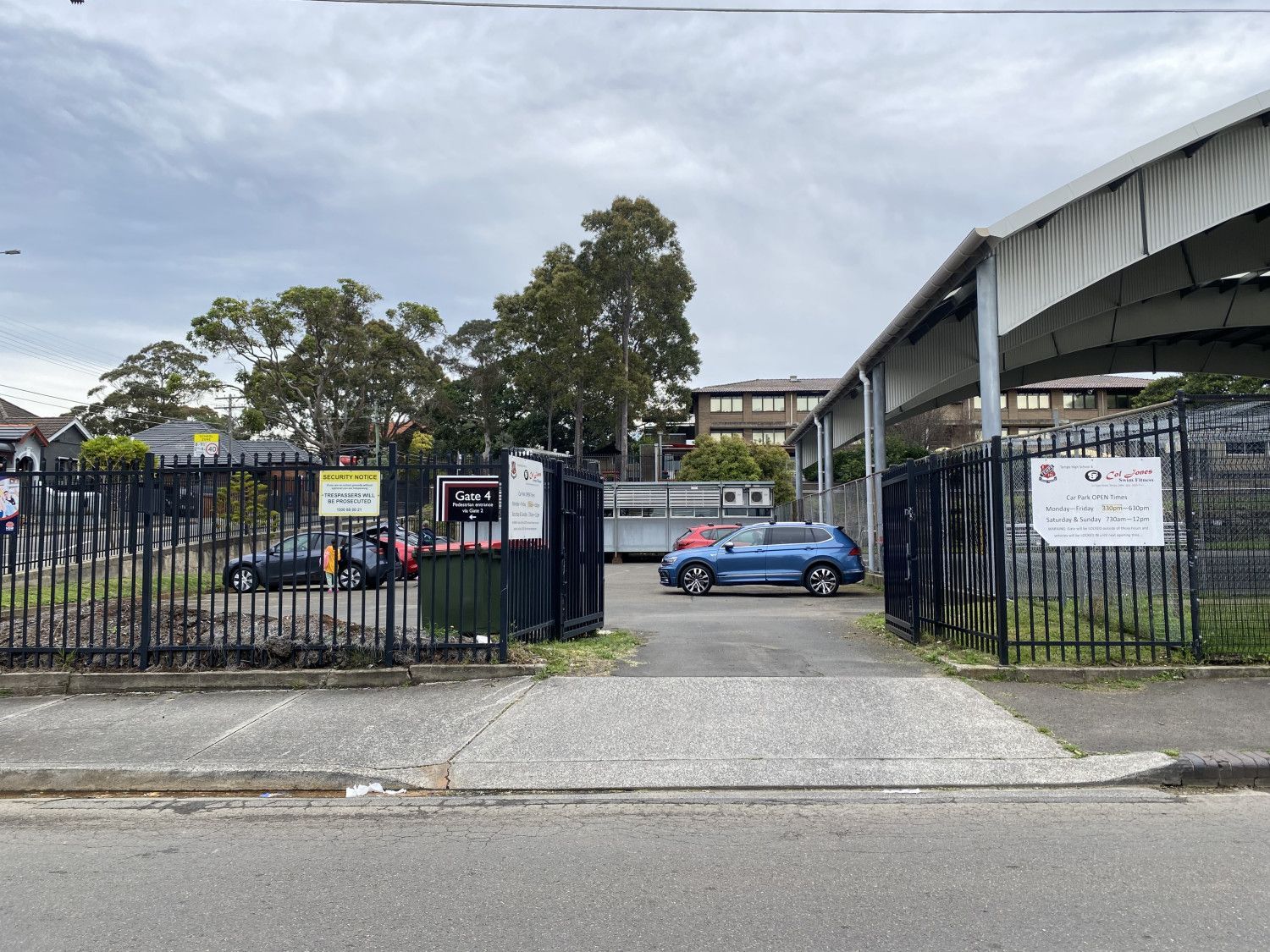 Open gate to a parking area with cars. Trees and buildings are visible under an overcast sky.