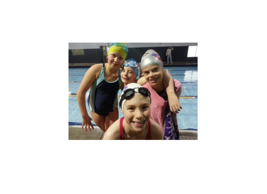 Four young people in swim caps and goggles pose by a pool, smiling.