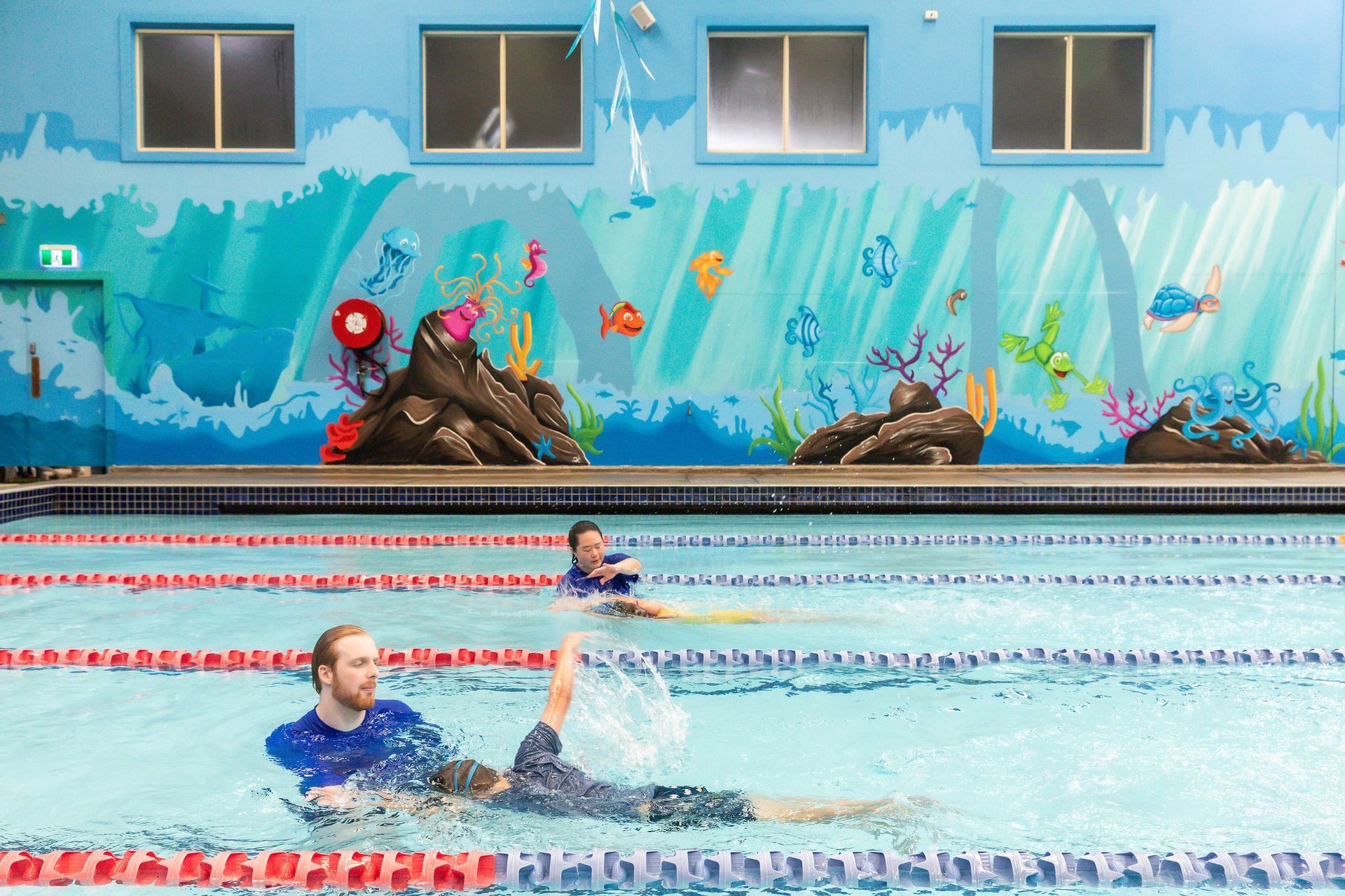 Two people swimming in a blue-walled indoor pool with an ocean-themed mural.