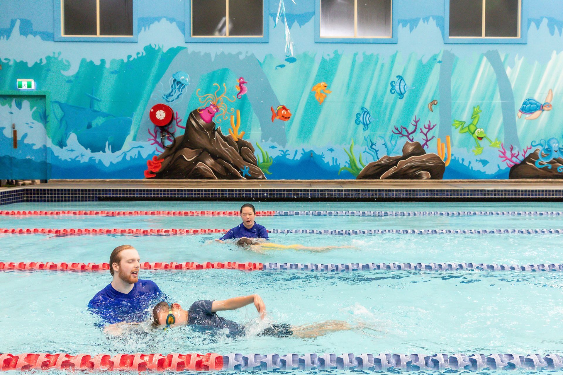 Swimmer in blue cap glides in an outdoor pool lane with mural-covered wall in the background