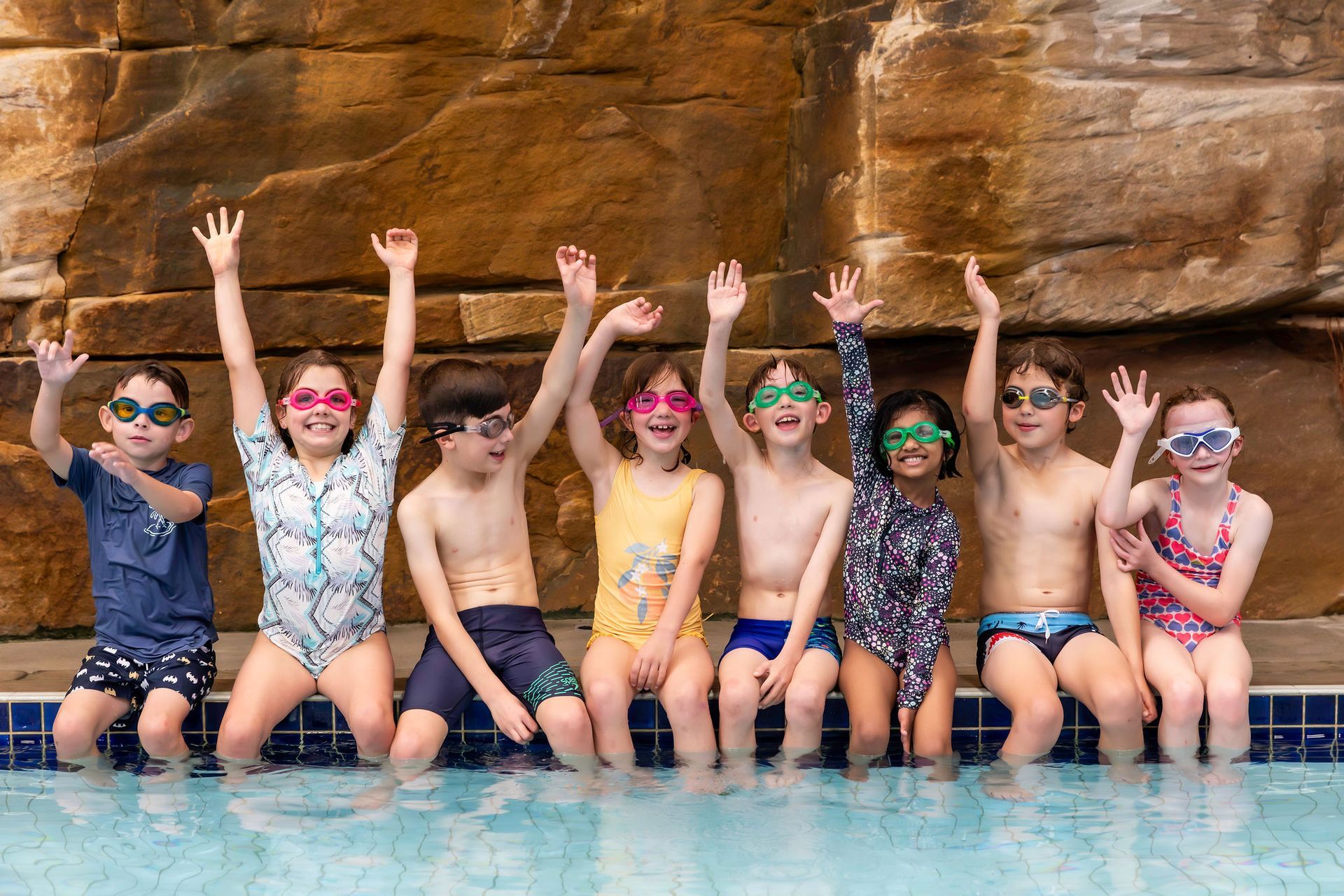 Eight children with goggles sit on a pool ledge against a stone wall, smiling and waving with hands raised.