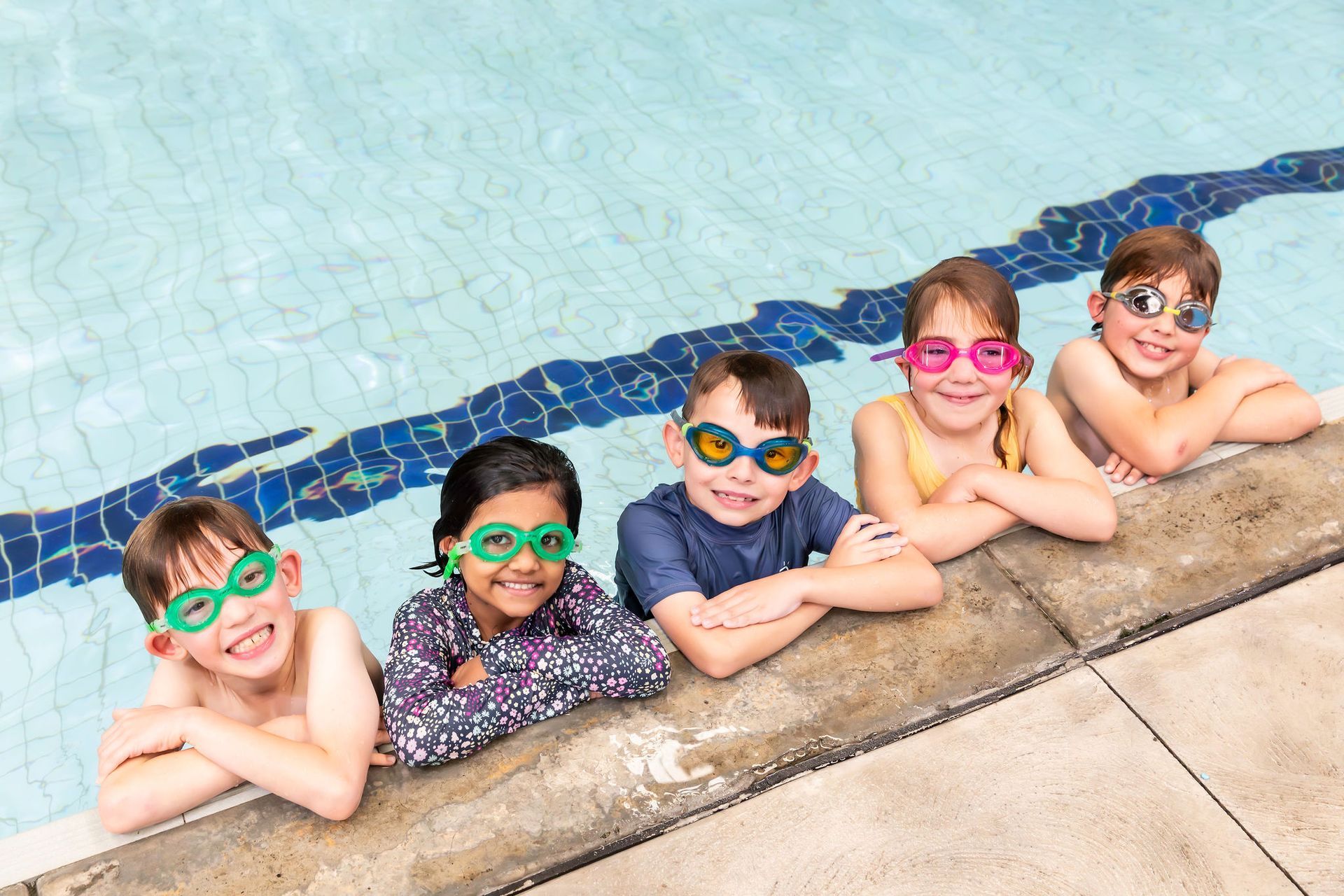 Five children in swim goggles smile at the edge of a pool.
