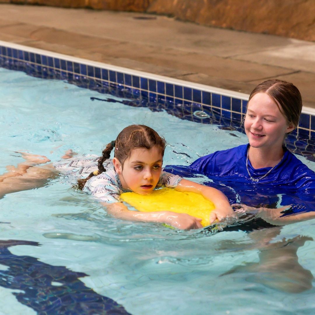 Girl in pool learning to swim with instructor using a yellow float.