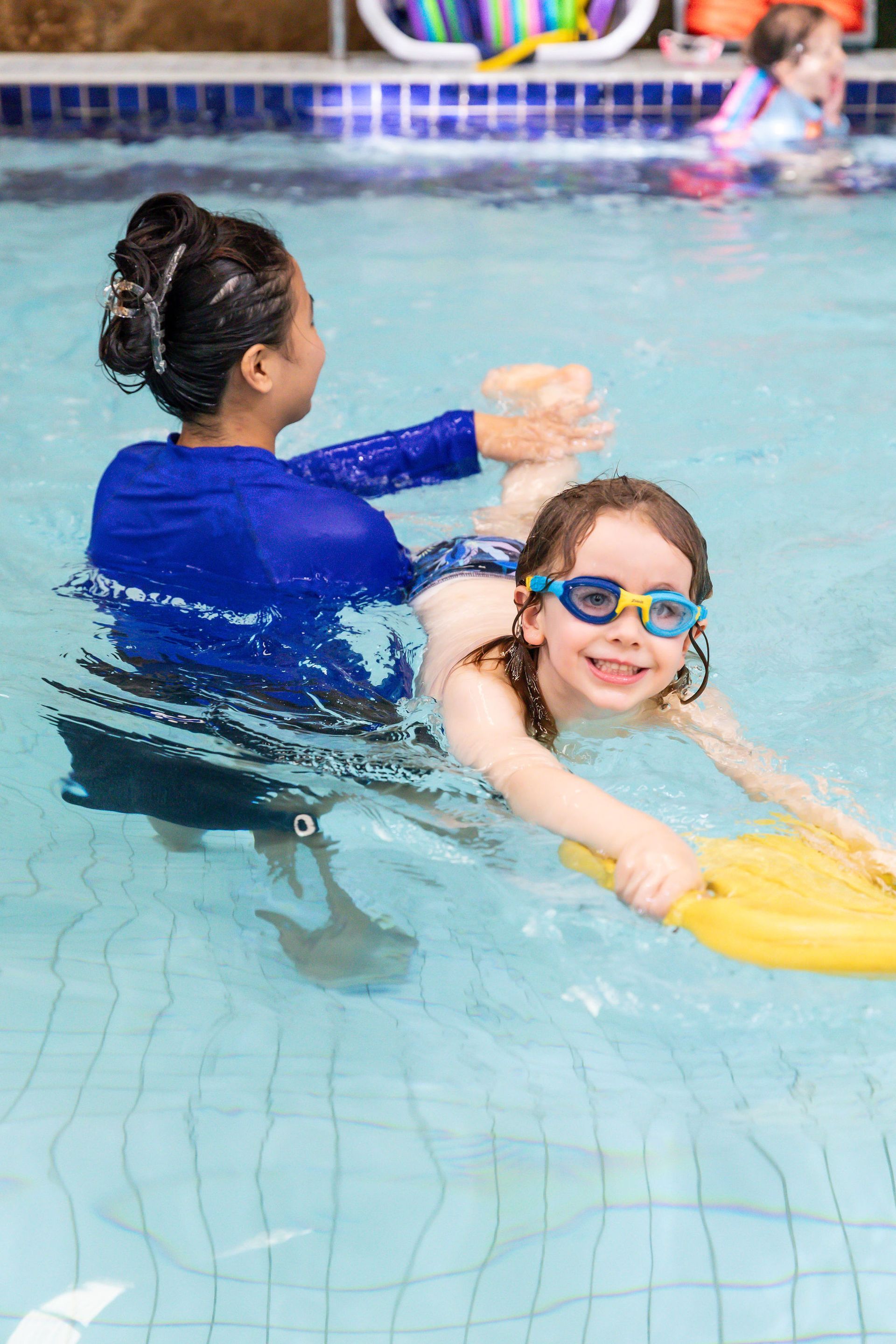 Child learning to swim with instructor in a pool, holding a kickboard.