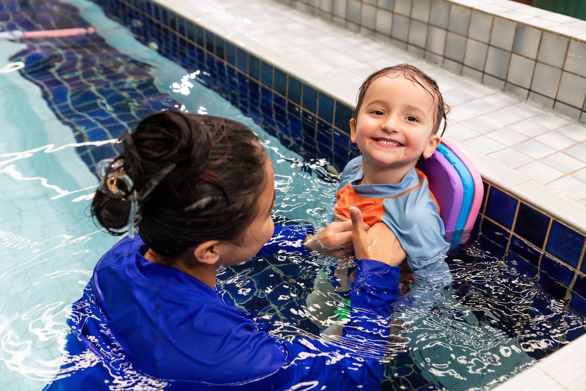 Adult assisting smiling child with confidence in a tiled pool during swimming lesson