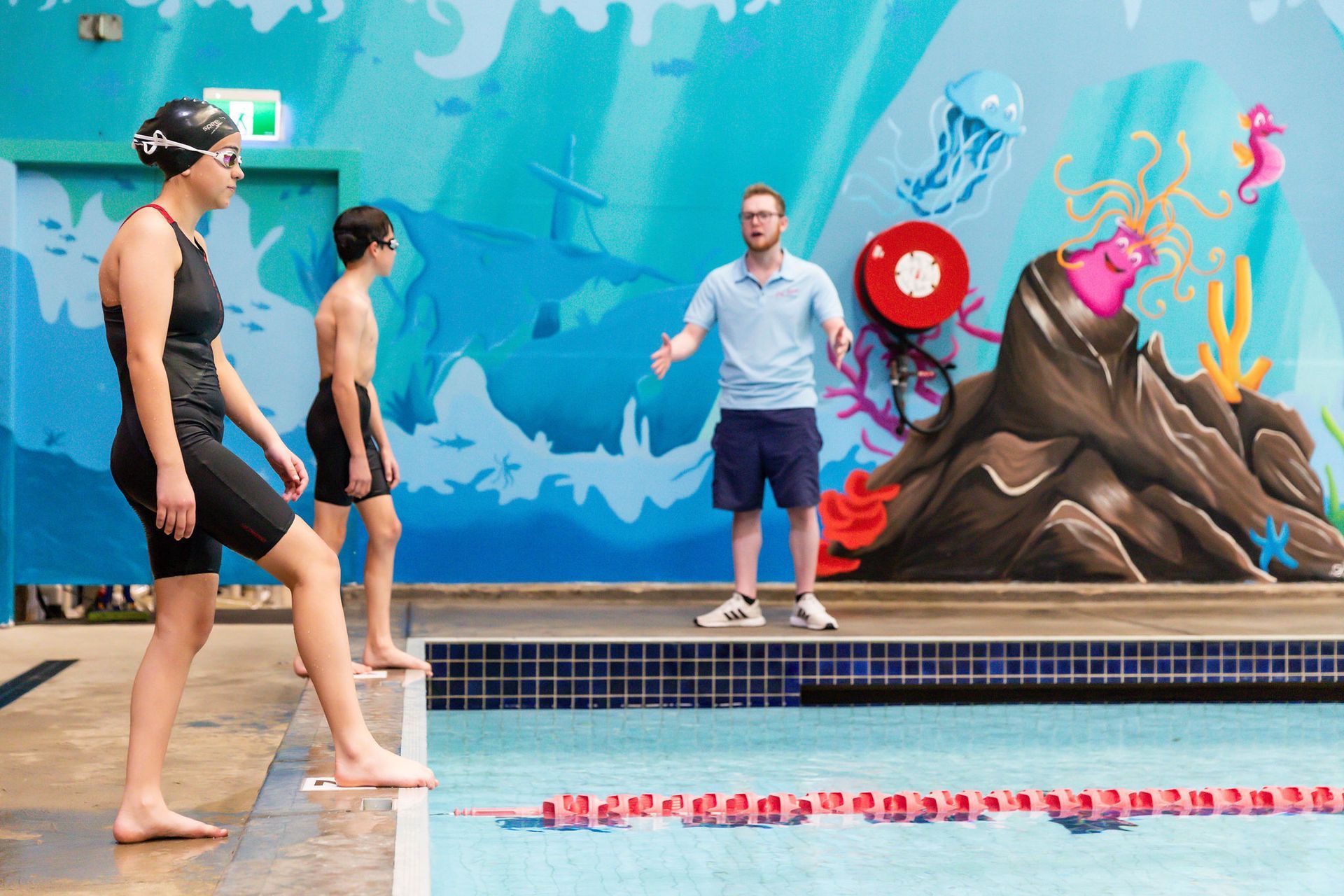 Swimmer and boy with coach at pool, ready to swim. Blue wall mural in background.