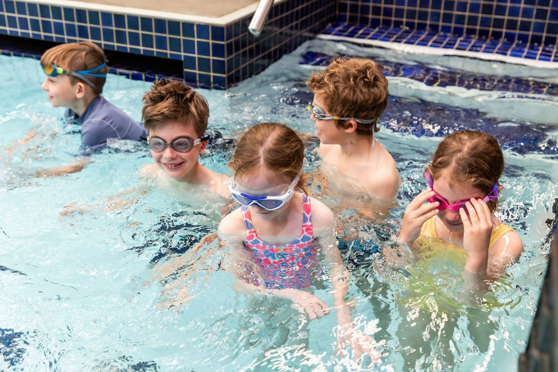 Children in a swimming pool, wearing goggles. They're splashing and smiling, enjoying a summer swim.