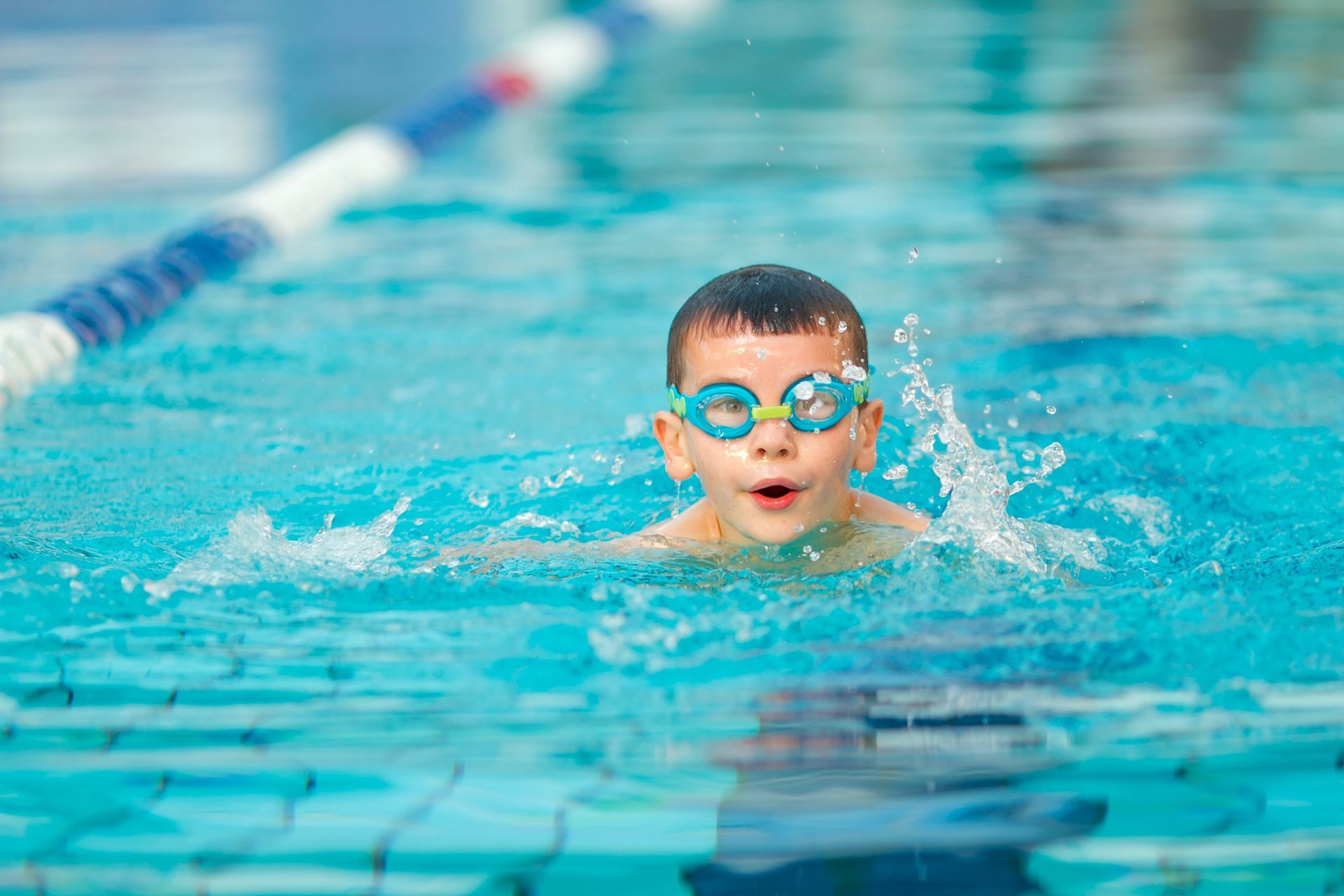 Boy in blue swim goggles swims in a pool with a surprised expression.