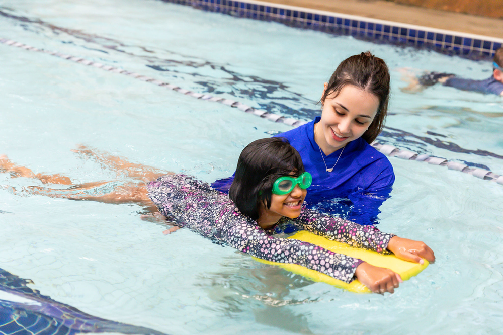 Child with goggles learning to swim in a pool with an instructor holding them from behind