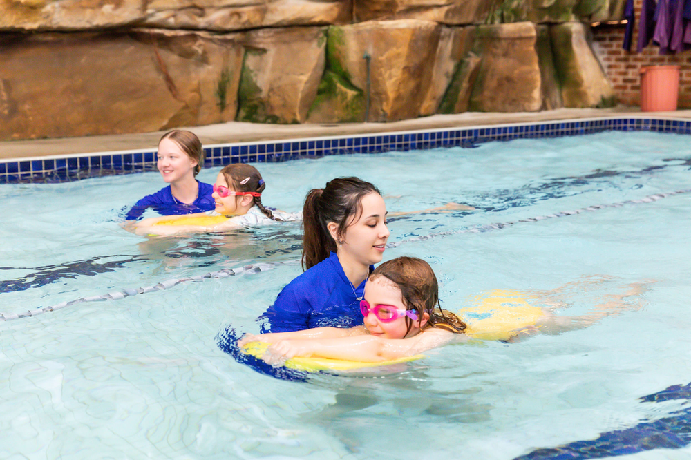 Swim instructor assisting a child in the pool with a yellow float, two other children nearby.