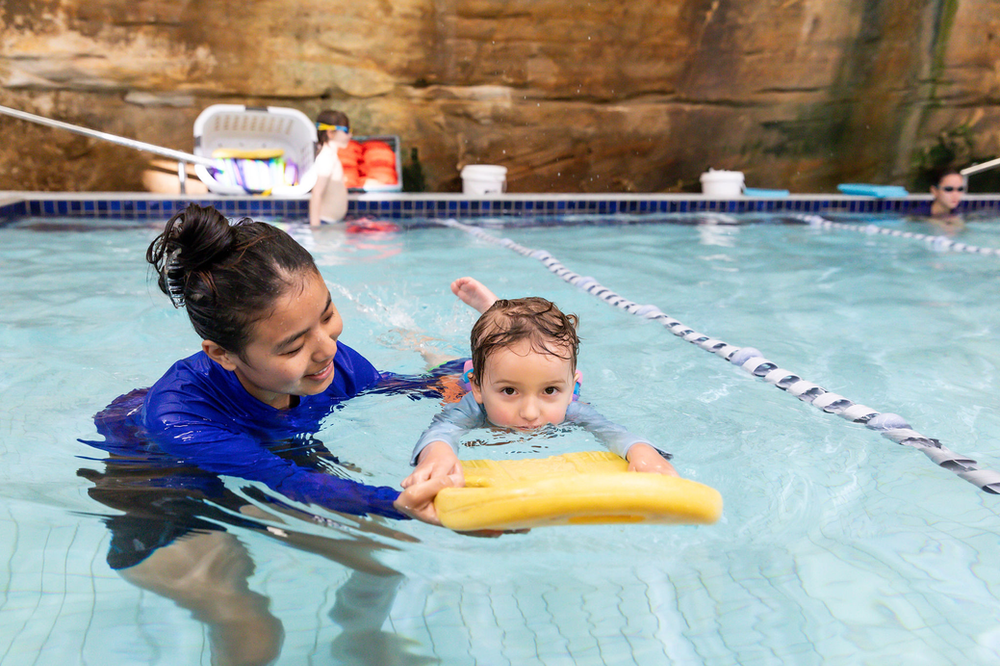 Person in blue shirt teaching a child to swim with a yellow kickboard in an indoor pool.