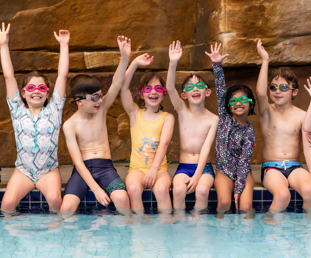 Group of kids in swim gear, goggles, arms raised, sitting at pool edge near a rock wall.